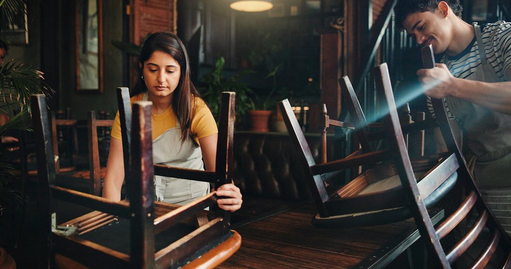 Dos personas colocan sillas boca abajo sobre las mesas de un restaurante; probablemente estén limpiando o cerrando al terminar la jornada. Una de ellas parece concentrada, mientras que la otra sonríe; el entorno cuenta con plantas y muebles de madera.