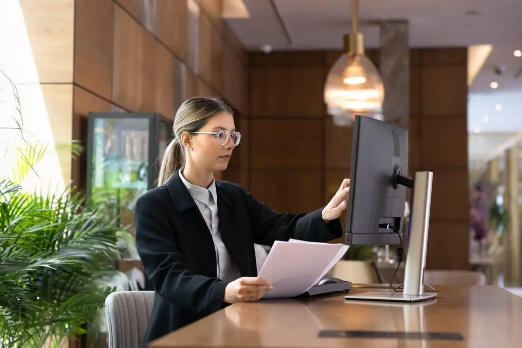 Una mujer con gafas y chaqueta de traje está de pie junto a un escritorio, sosteniendo unos papeles con una mano y ajustando la pantalla del ordenador con la otra, en un entorno de oficina moderno.