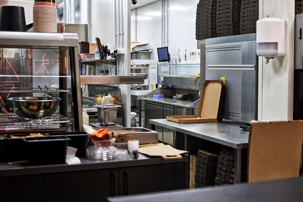 Una cocina industrial con encimeras de acero inoxidable, cajas de pizza apiladas, cajas de pizza abiertas, utensilios y equipamiento de cocina. El espacio parece limpio y ordenado, listo para la preparación de alimentos.