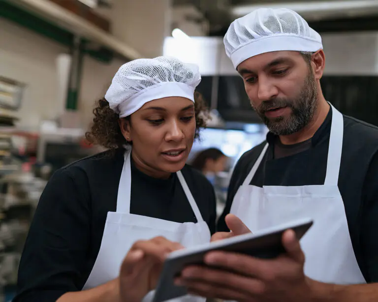 Dos panaderos, con delantales blancos y redecillas en el pelo, miran juntos una tableta digital en una cocina industrial; parecen concentrados y enfrascados en una conversación.