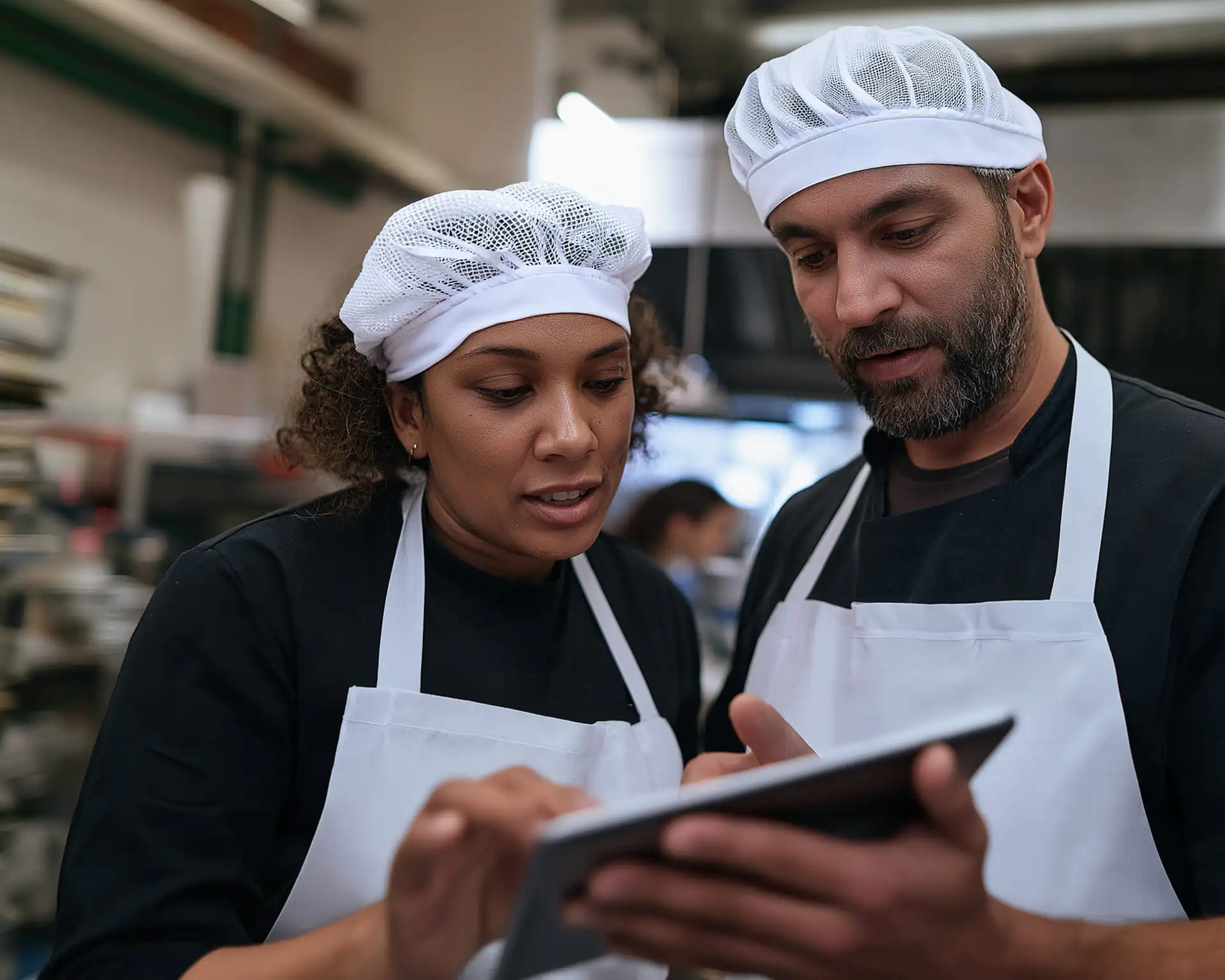 Dos panaderos, con delantales blancos y redecillas en el pelo, miran juntos una tableta digital en una cocina industrial; parecen concentrados y enfrascados en una conversación.