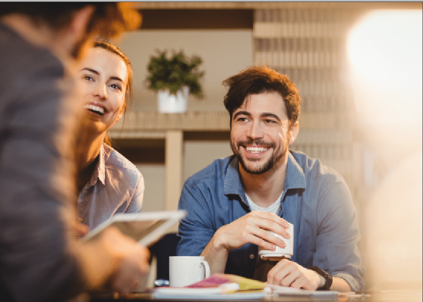 Trois personnes sont assises à une table, souriantes et en pleine conversation. L'une d'elles tient une tasse, une autre tient une tablette, et il y a des livres ainsi qu'une tasse à café sur la table. Une plante en pot se trouve à l'arrière-plan.