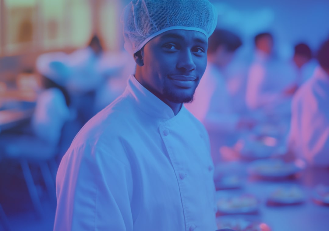 Un chef, vêtu d'un uniforme blanc et d'une charlotte, sourit à la caméra dans une cuisine où d'autres chefs se trouvent en arrière-plan, sous un éclairage bleu et violet.