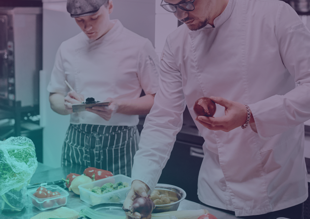 Deux chefs en tenue blanche se tiennent dans une cuisine. L'un d'eux prend des notes sur un bloc-notes, tandis que l'autre tient un oignon rouge au-dessus d'un plan de travail sur lequel sont disposés des légumes, de la laitue, des tomates, des raisins et d'autres ingrédients.