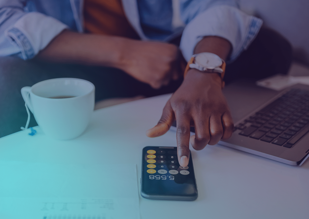 Une personne utilise une application de calculatrice sur son smartphone, à côté d'un ordinateur portable et d'une tasse de café au lait posés sur une table ; elle semble s'occuper de ses finances ou de son budget.