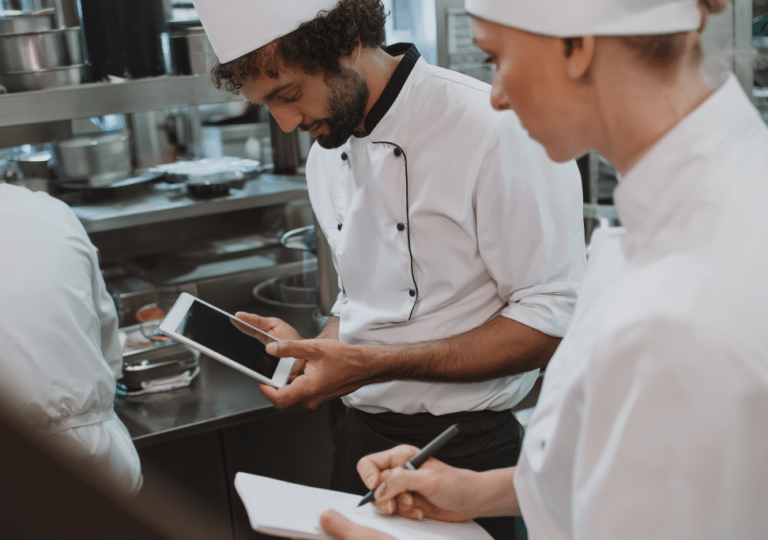 Deux chefs, vêtus d'uniformes blancs et coiffés de toques, travaillent dans une cuisine ; l'un prend des notes sur un bloc-notes tandis que l'autre utilise une tablette. On aperçoit à l'arrière-plan du matériel et des ustensiles en inox.