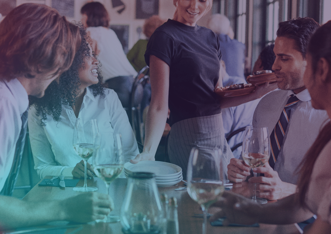 Quatre personnes sont assises à une table de restaurant, un verre de vin à la main, en train de discuter et de sourire, tandis qu'un serveur tend un plat à l'une d'entre elles. L'ambiance est animée et conviviale.
