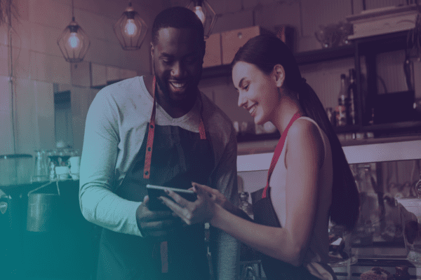 Deux personnes souriantes, vêtues de tabliers, se tiennent dans un café ou une cuisine chaleureux, les yeux rivés ensemble sur une tablette, sous des suspensions lumineuses qui diffusent une lumière chaleureuse.
