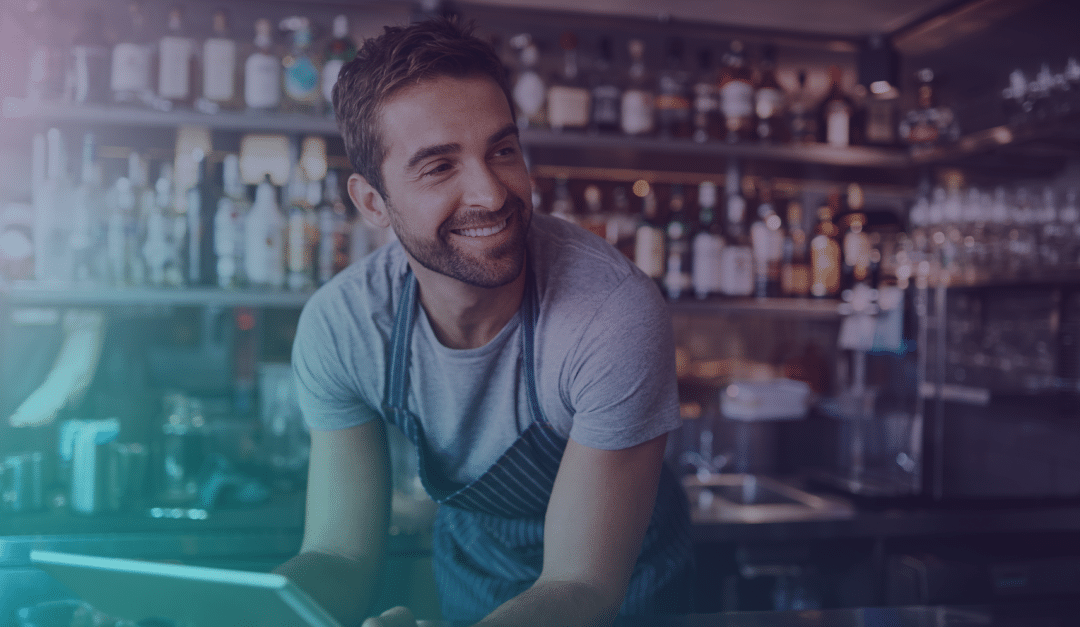 Un barman souriant, vêtu d'une chemise grise et d'un tablier, se tient derrière un comptoir, entouré de bouteilles et d'ustensiles de bar, le regard tourné vers le côté.