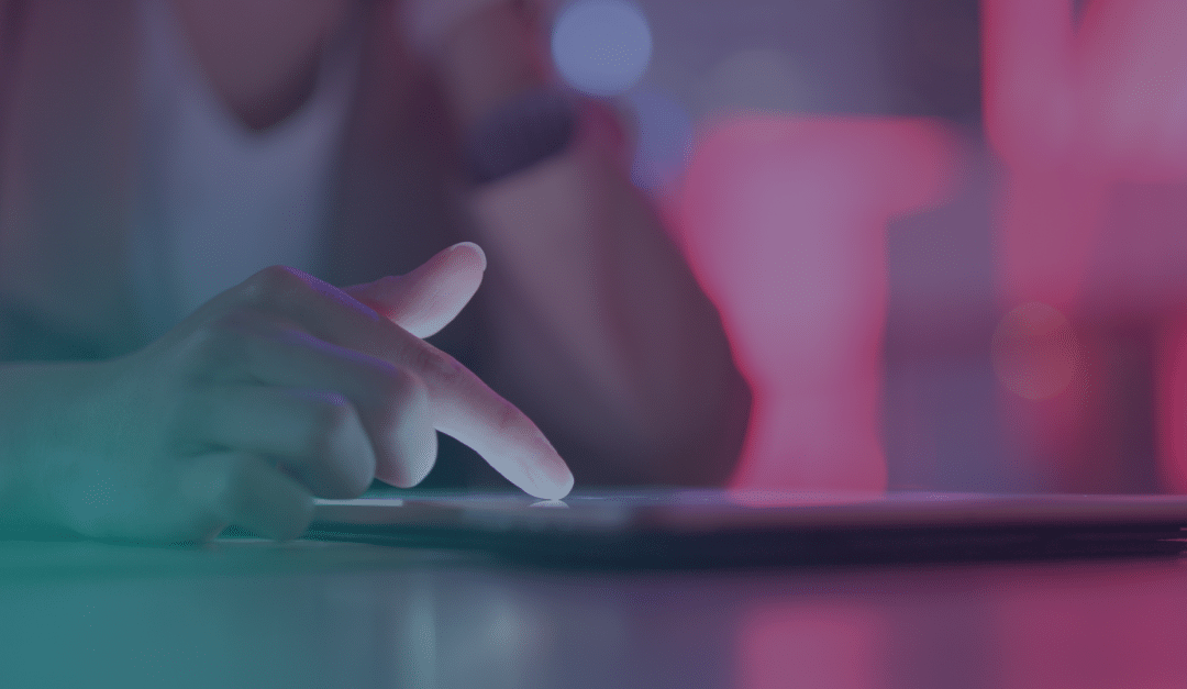 La main d'une personne effleure l'écran d'une tablette dans une pièce faiblement éclairée, baignée d'une lumière rose et bleue, créant ainsi une ambiance moderne et axée sur la technologie.