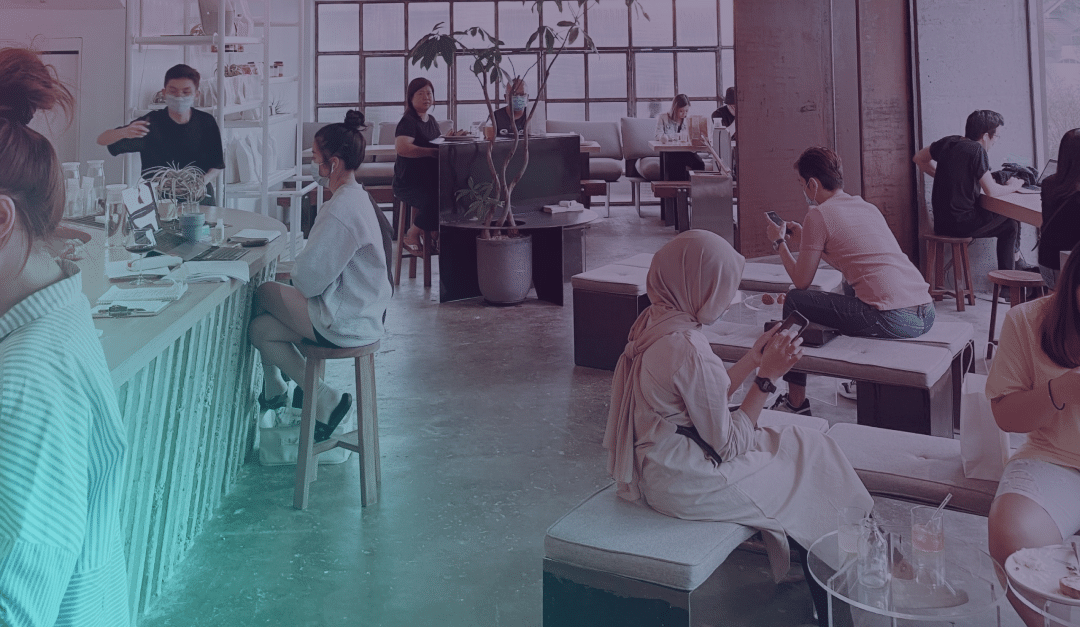 Les clients sont assis et discutent dans un café moderne et lumineux, doté de grandes baies vitrées. Certains travaillent sur leur ordinateur portable, tandis que d'autres discutent ou consultent leur téléphone. Un barista prépare des boissons derrière le comptoir.