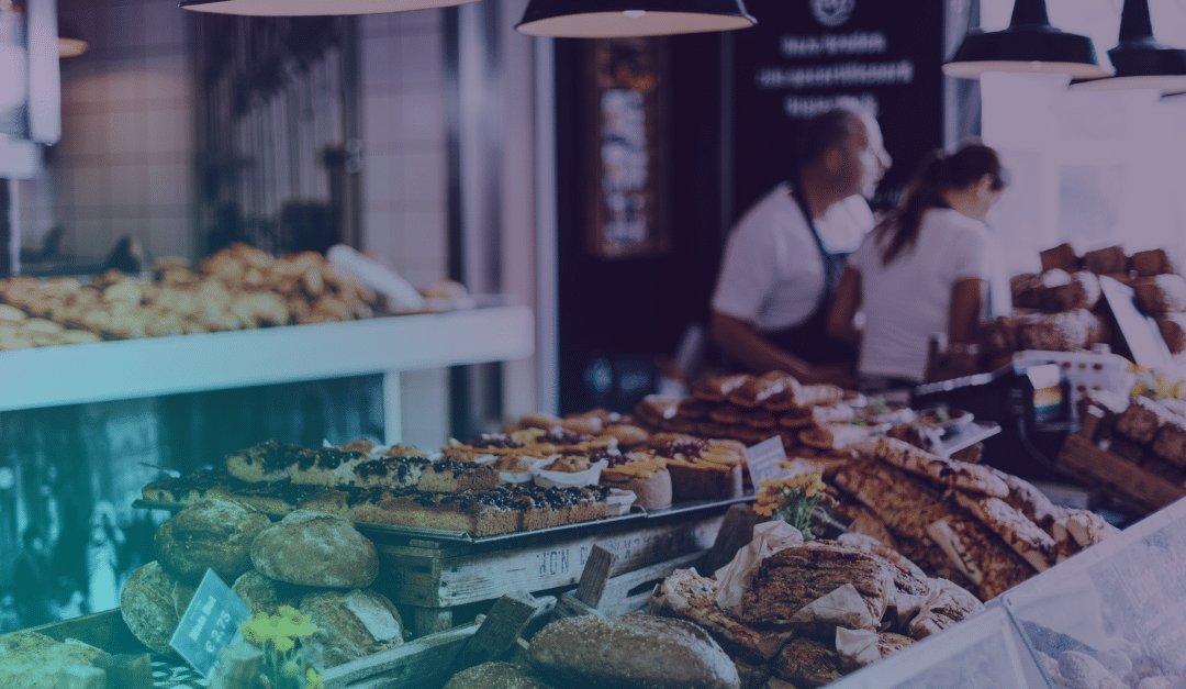 Une boulangerie présente toute une gamme de pains et de viennoiseries, tandis que deux personnes discutent derrière le comptoir. La scène est baignée d'une lumière chaleureuse et accueillante.