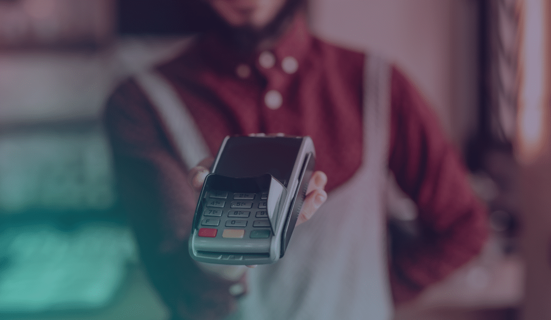 Une personne vêtue d'un t-shirt rouge et d'un tablier tient un terminal de paiement face à la caméra, invitant à effectuer un paiement sans contact. L'arrière-plan est flou, ce qui permet de mettre l'accent sur le lecteur de carte.