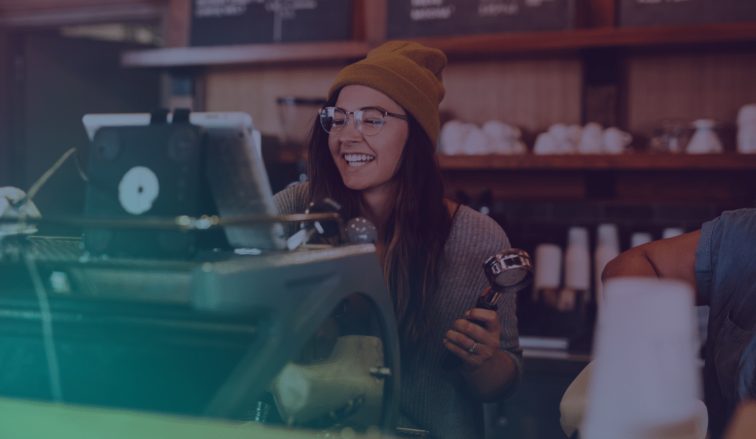 Un barista, portant des lunettes et un bonnet, sourit tout en tenant un porte-filtre derrière une machine à café dans un café.