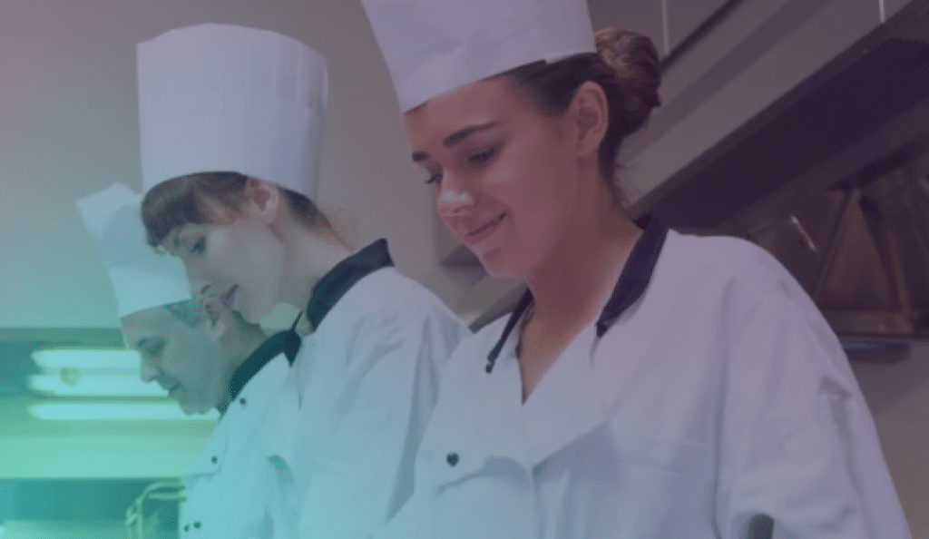 Trois chefs, vêtus d'uniformes blancs et coiffés de toques, sont alignés dans une cuisine, concentrés sur leur travail.