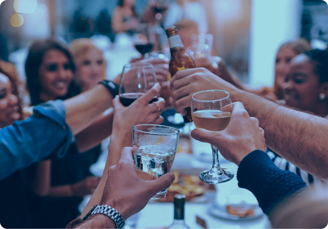 Un groupe de personnes assises autour d'une table font tinter leurs verres et leurs bouteilles pour trinquer, dans le cadre d'une fête animée.