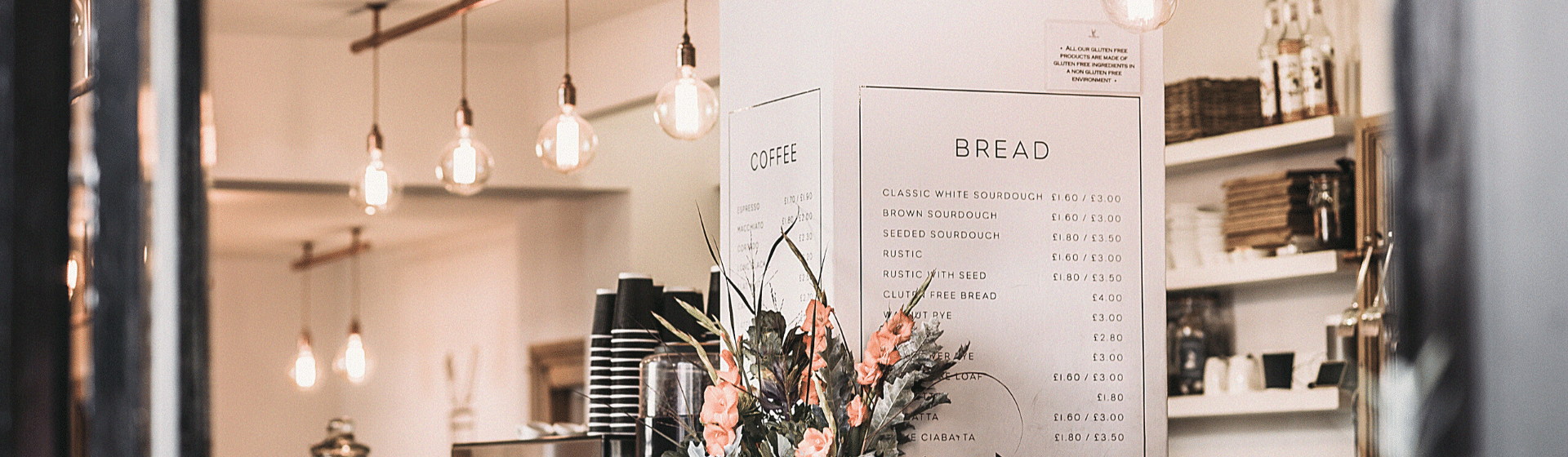 L'intérieur chaleureux d'un café, avec des ampoules suspendues, des étagères garnies de bocaux et de tasses, un bouquet de fleurs et, au centre, un tableau présentant la carte proposant différents cafés et pains.