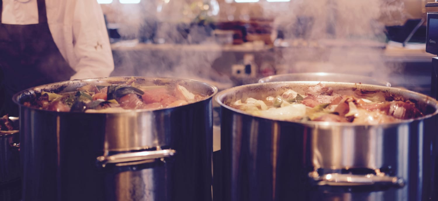 Deux grandes casseroles en inox remplies de légumes et de bouillon mijotant, posées sur une cuisinière dans une cuisine animée. Une personne vêtue d'une veste de chef se tient à proximité, légèrement floue à l'arrière-plan.