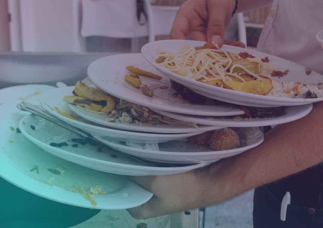 Une personne tient à deux mains une grande pile d'assiettes sales sur lesquelles se trouvent des restes de nourriture, notamment des spaghettis, des haricots et des légumes. Les assiettes sont en train d'être débarrassées, probablement après un repas ou dans un restaurant.