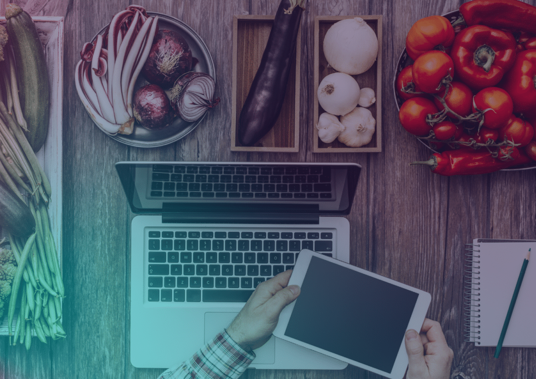 Une personne tenant une tablette est assise devant un ordinateur portable, entourée de légumes frais, notamment des tomates, des champignons, des aubergines et des oignons verts ; un cahier et un stylo se trouvent à proximité, posés sur une table en bois.