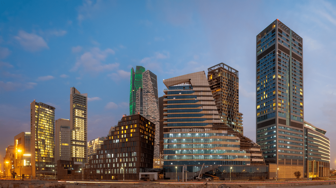 A cityscape at dusk shows modern skyscrapers with unique, angular designs. Buildings are lit from within and the sky is filled with soft clouds, transitioning from blue to light orange.