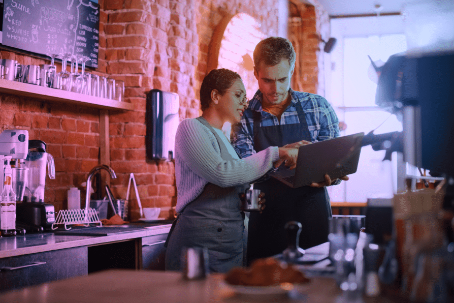 Two café workers in aprons stand behind the counter, looking at a laptop together. They appear to be discussing something, surrounded by coffee equipment and exposed brick walls.