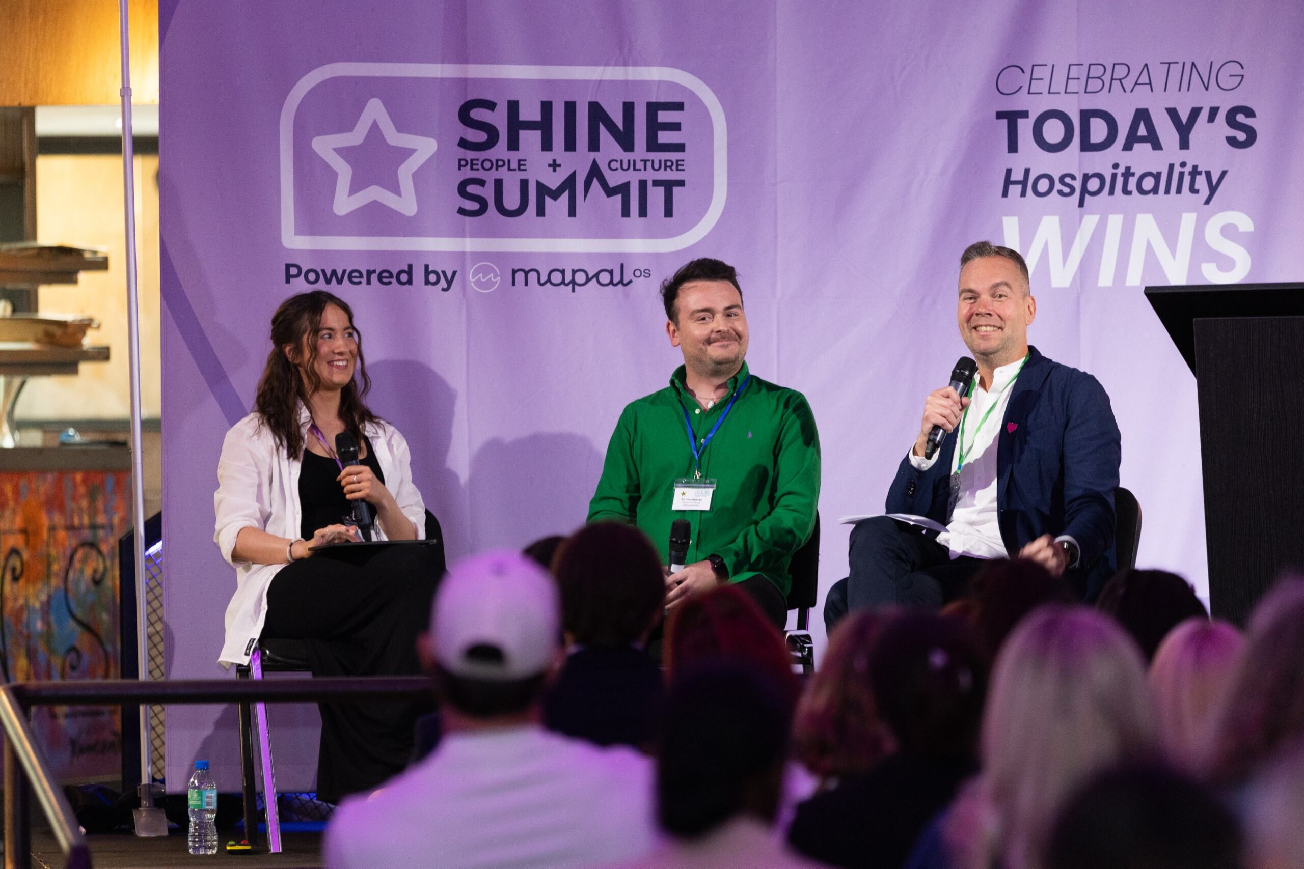 Three people sit on stage holding microphones, speaking at the SHINE People + Culture Summit in front of an audience, with a large purple event banner in the background.