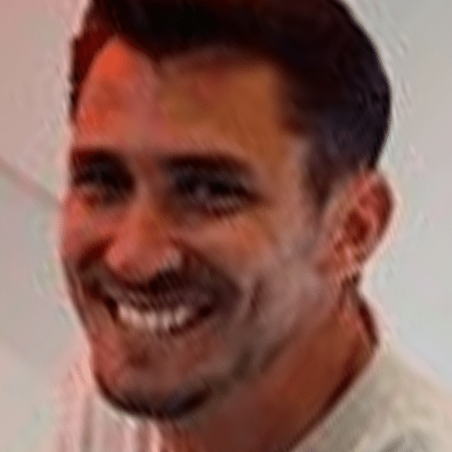 A man with short dark hair and facial hair smiles at the camera, wearing a light-coloured shirt, with a blurred light background.