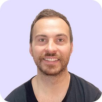 A smiling man with short brown hair and a trimmed beard is wearing a black shirt. The background is light purple with rounded corners.