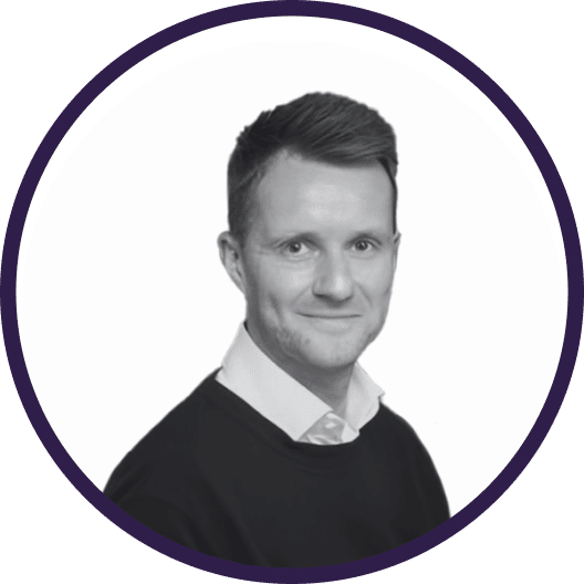 A man with short hair, wearing a dark jumper over a collared shirt, smiles at the camera. The image is black and white and framed by a circular purple border.