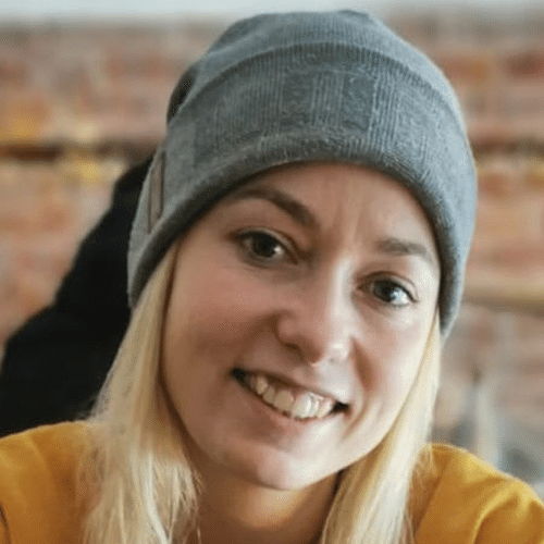 A woman with long blonde hair wearing a grey beanie and a yellow top smiles at the camera. The background is softly blurred with exposed brick visible.