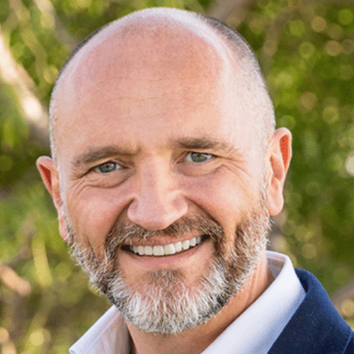 Smiling middle-aged man with a bald head and salt-and-pepper beard, wearing a white shirt and dark jacket, standing outdoors with green foliage in the background.
