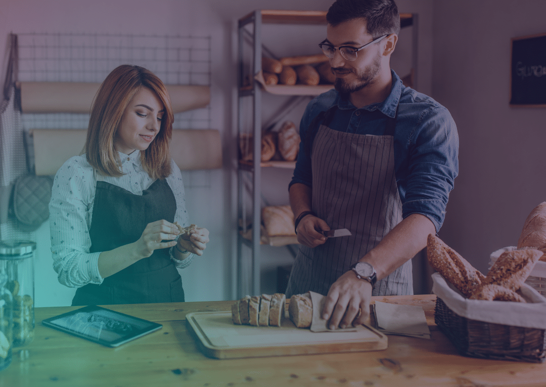 Two people wearing aprons stand at a bakery counter. One is slicing bread on a chopping board while the other looks at her hands. There are baguettes in a basket and a tablet on the wooden counter.