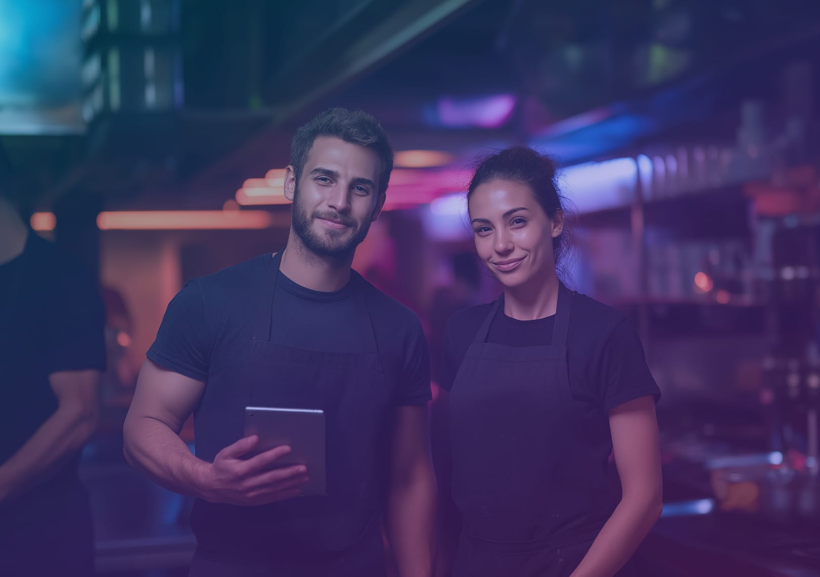 Two restaurant staff members in black aprons stand side by side in a kitchen, smiling at the camera. One holds a tablet. The background shows kitchen equipment and colourful lighting.