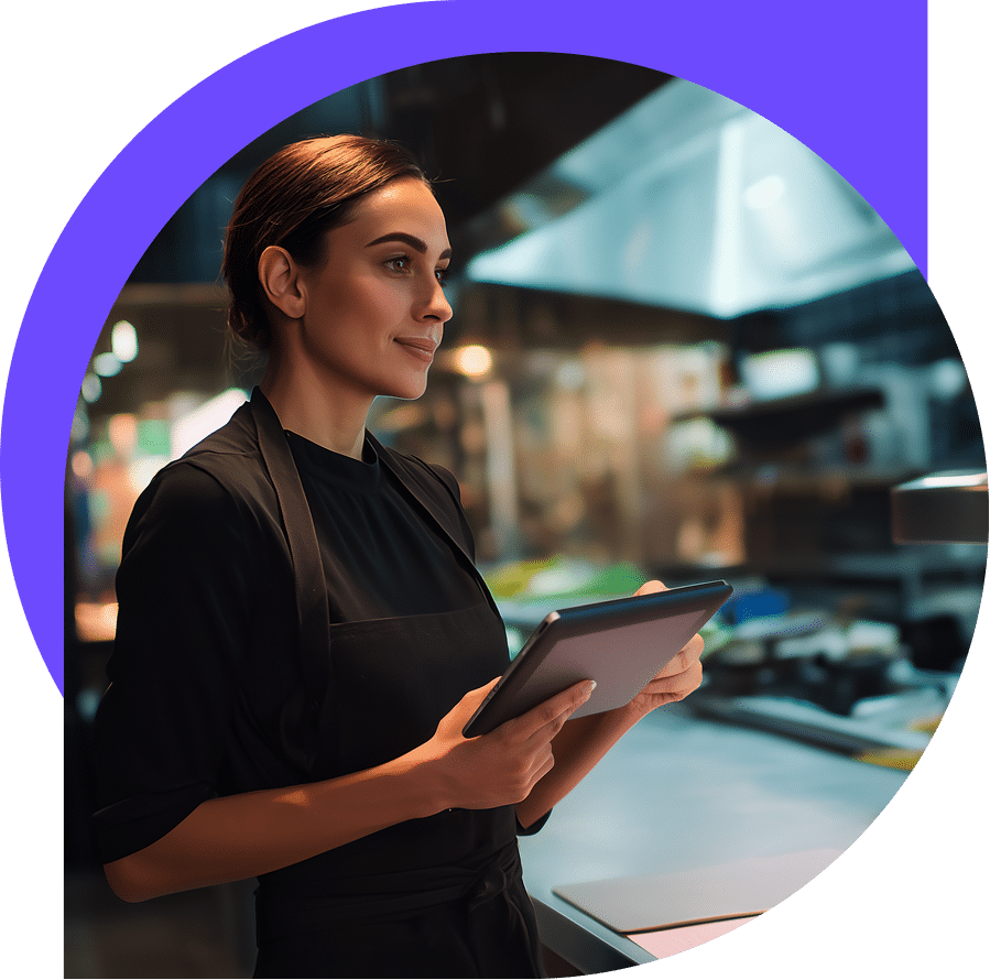 A woman in a black apron holds a tablet and stands in a modern, well-lit kitchen, appearing focused and thoughtful.