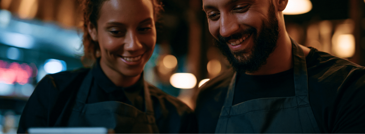 Two people wearing aprons smile and look at a screen together in a warmly lit indoor setting, possibly a restaurant or café.