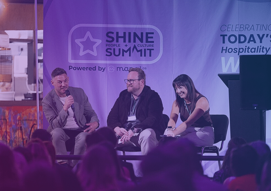 Three speakers sit on stage in front of a Shine Summit banner, addressing an audience. One man is speaking into a microphone, whilst the others listen and smile. A lectern stands nearby.