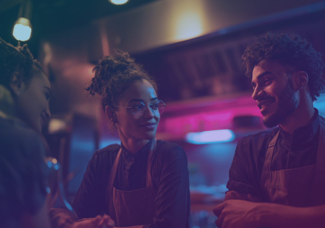 Three people wearing aprons sit and converse in a warmly lit, modern kitchen with colourful lighting, suggesting a relaxed and friendly atmosphere, possibly at work or during a break.