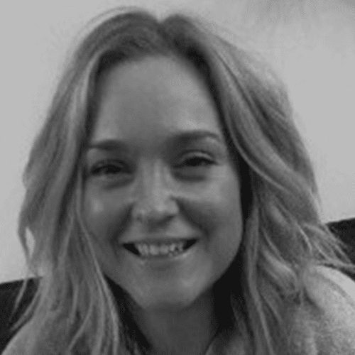 A woman with wavy, shoulder-length hair smiles at the camera. The photo is in black and white, and she is sitting indoors with a light background.