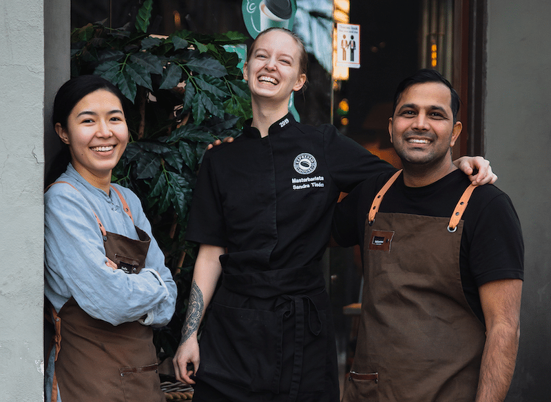 Three people in work uniforms stand outside a building, smiling and posing together. One is wearing a black chef's coat, while the others wear brown aprons. There is a green plant in the background.