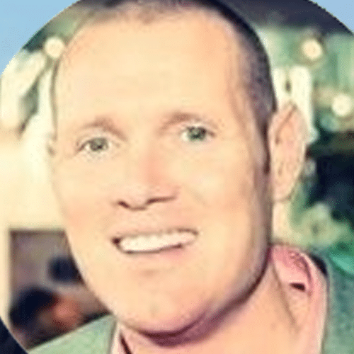 A man with short hair is smiling at the camera, wearing a light-coloured shirt and jacket. The background is blurred, suggesting an indoor setting.