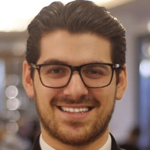 A man with dark hair and glasses smiles at the camera. He has a trimmed beard and is wearing a dark suit jacket over a white shirt. The background is softly blurred with warm lighting.