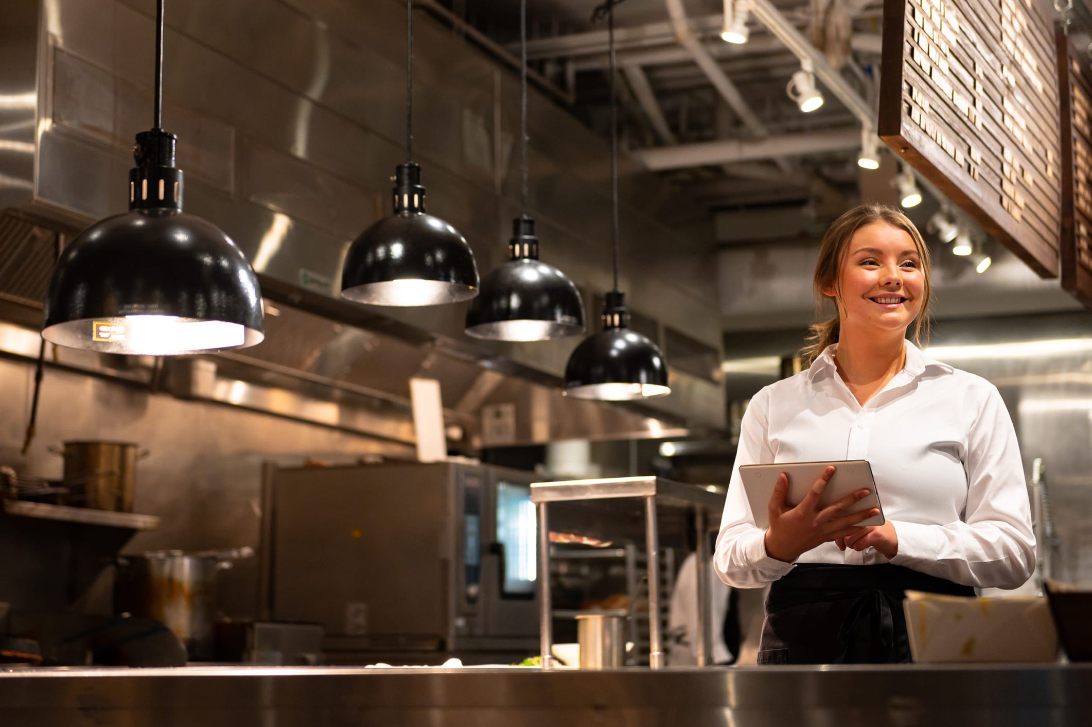 A woman in a white shirt stands in a modern restaurant kitchen, smiling and holding a digital tablet under industrial hanging lights.