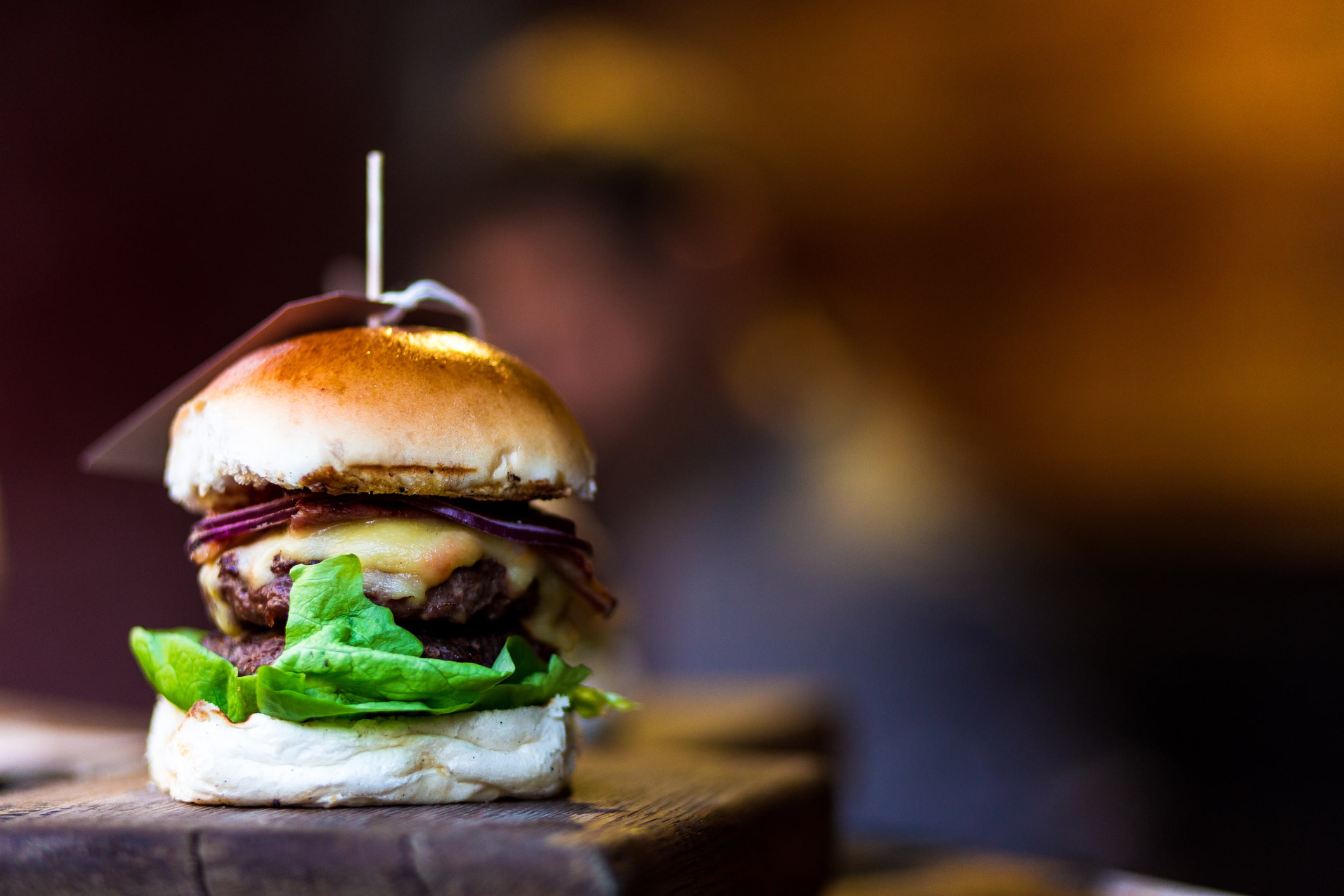 A close-up of a cheeseburger with lettuce, red onion, and a beef burger, held together with a cocktail stick, on a wooden surface with a blurred background.