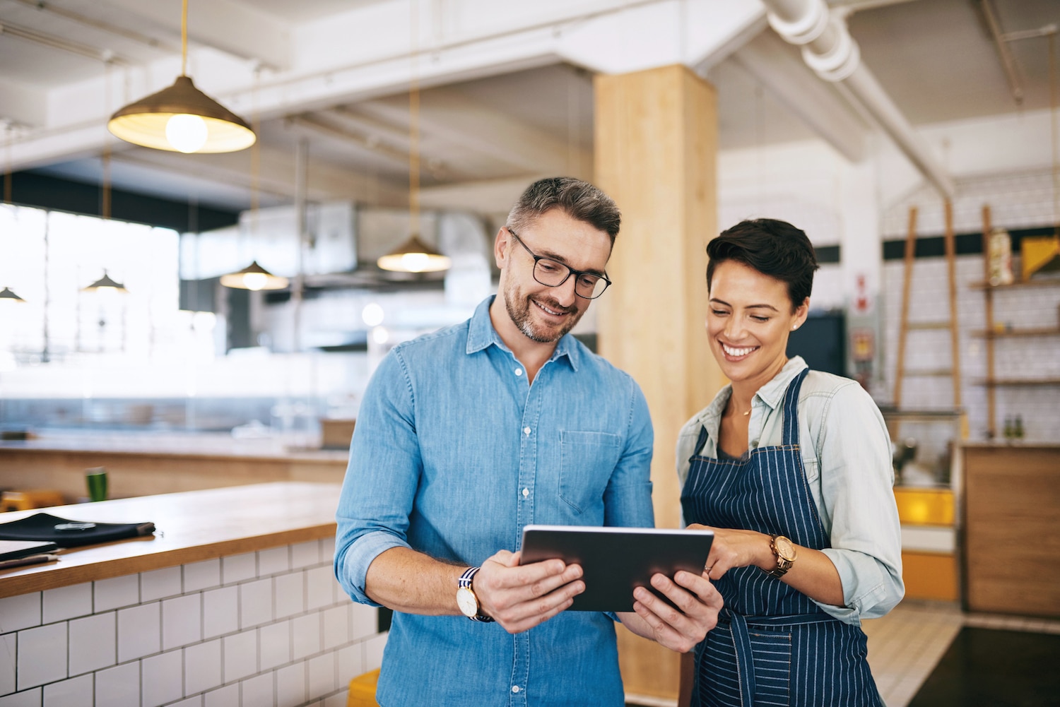 Two people stand in a modern café, smiling and looking at a tablet together. One wears glasses and a denim shirt, the other wears an apron. Bright lights and wooden décor are visible in the background.