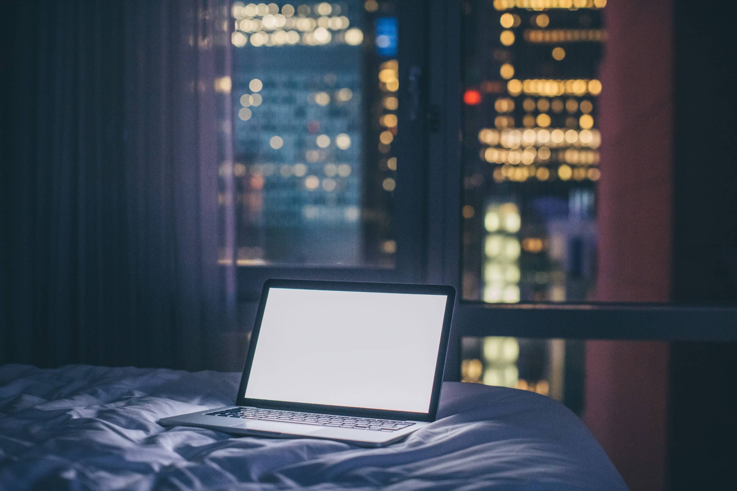 A laptop with a bright, blank white screen sits on a bed at night, with a window showing blurred city lights in the background.