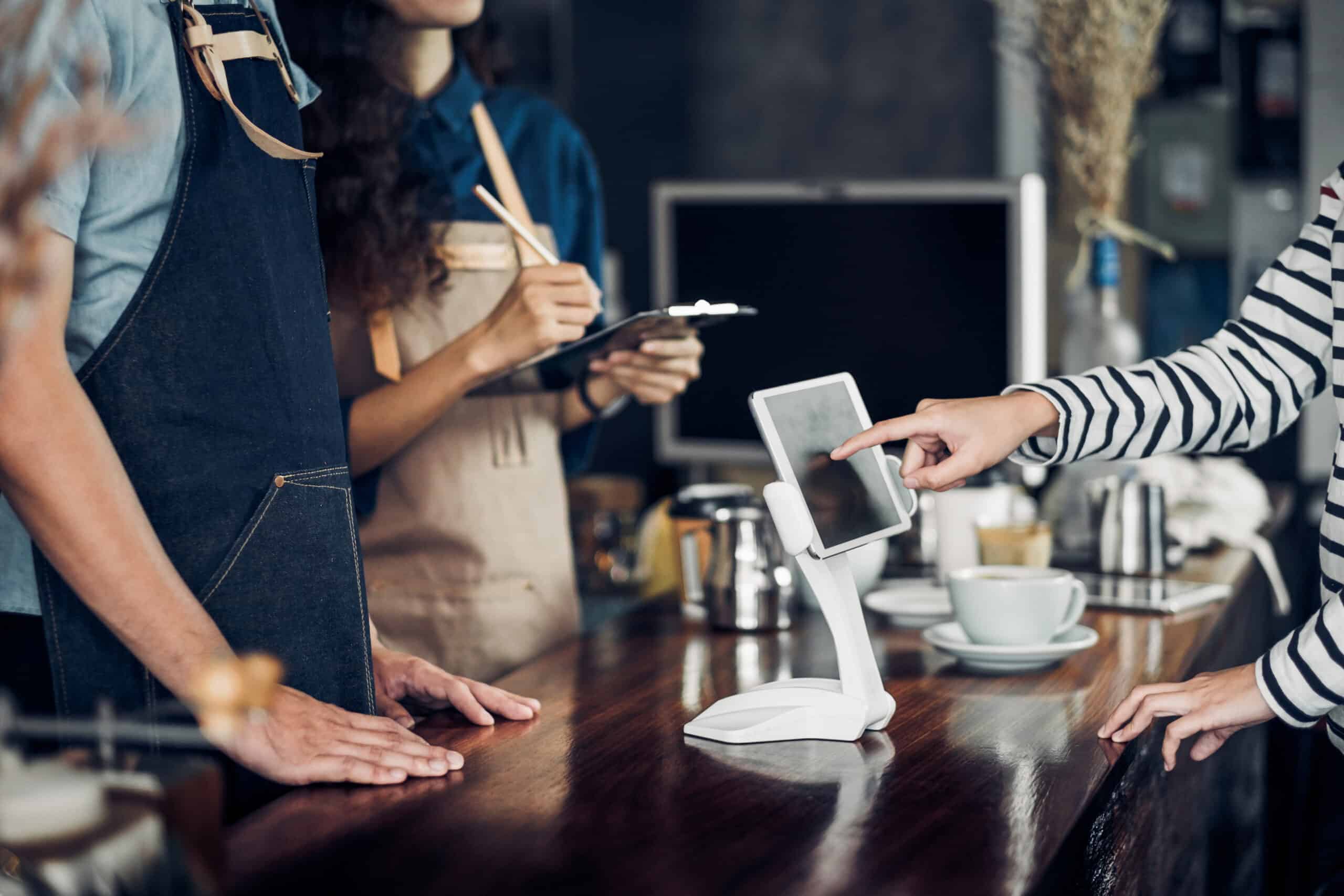 A person touches a digital payment screen on a café counter while two café staff in aprons stand nearby, one holding a clipboard. Coffee cups and saucers are on the wooden counter.