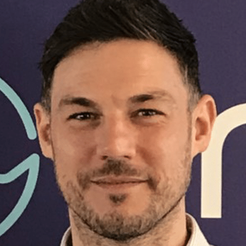 A man with short dark hair and light facial hair is smiling slightly in front of a purple background with light blue and white designs.