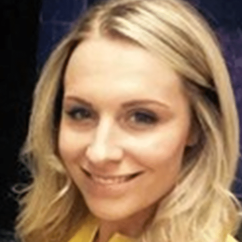 A woman with long blonde hair smiles at the camera. She is wearing a yellow top and is in front of a dark, blurred background.