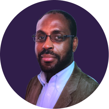 A man with short hair, a beard, and glasses, wearing a brown jacket over a white shirt, poses in front of a solid dark purple background.